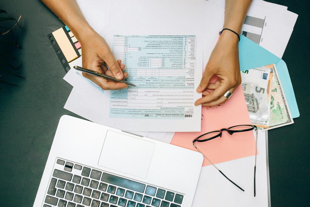 Hands writing on tax documents with laptop, glasses, and currency on desk.