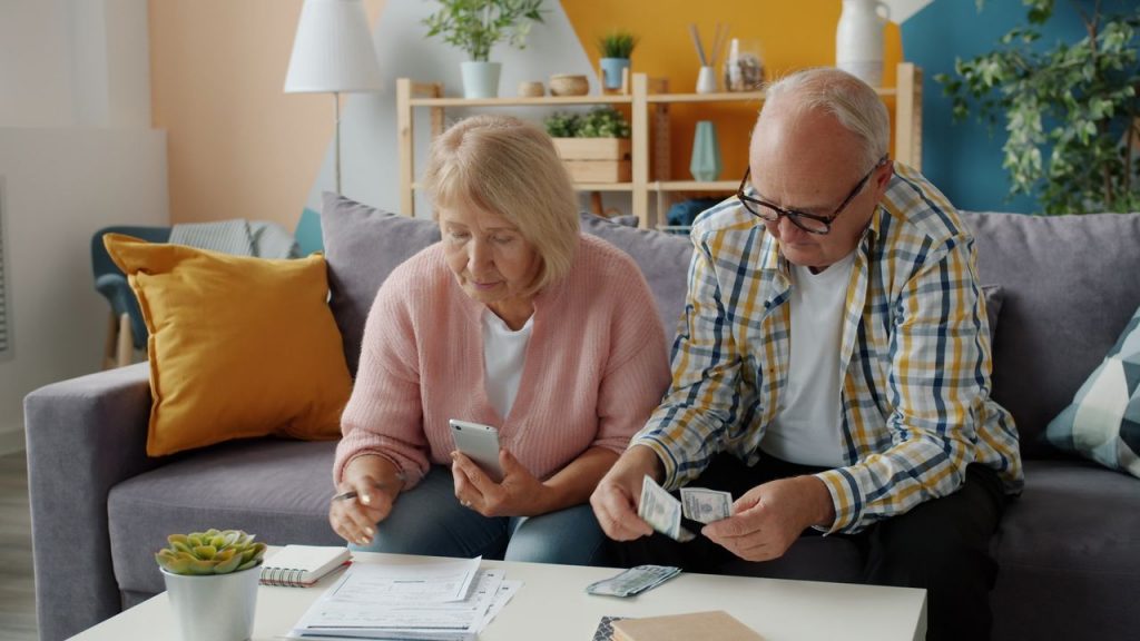 Elderly couple budgeting at home, seated on a cozy sofa, using smartphone and cash.